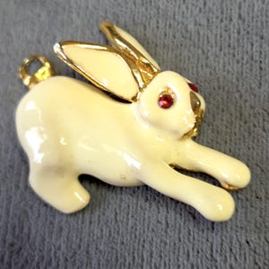 May include: A vintage-style bunny brooch in cream enamel with gold-tone accents. The bunny has red rhinestone eyes and is posed in a running position. The brooch has a pin closure on the back.