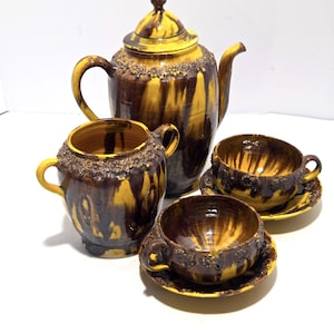 May include: A ceramic tea set with a teapot, sugar bowl, and two teacups with saucers. The set features a brown and yellow glaze with a textured floral design around the rims. The teapot is approximately 10 inches tall.