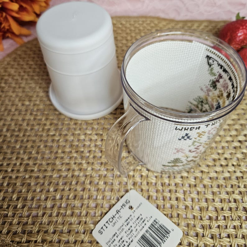Pu&ograve; includere: Una tazza di plastica trasparente con un inserto in tessuto bianco che dice "When This Mug Is Empty" in ricamo nero. La tazza &egrave; appoggiata su una superficie intrecciata marrone con un coperchio di plastica bianco e un'etichetta bianca che dice "Stitch-A-Mug" in nero.