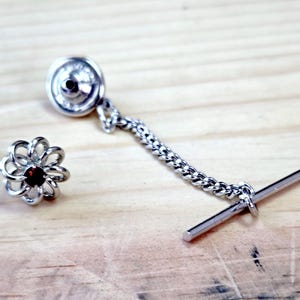 May include: A silver-toned lapel pin featuring a circular, open-work flower design with a small, dark red gemstone at its center. The pin includes a short, delicate metal chain attached to a straight, horizontal bar fastener at the other end.