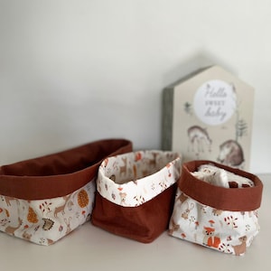Peut inclure: Trois paniers de rangement en tissu avec un fond blanc et un motif de cerf, de renard et de hérisson bruns. Les paniers ont une doublure en tissu brun et sont disposés en rangée sur une surface blanche.