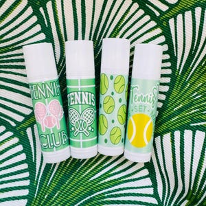 May include: Four white lip balm tubes with green and white tennis-themed designs. The tubes have different designs, including tennis rackets, tennis balls, and the words "Tennis Club" and "Tennis Set Match".