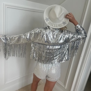 A woman in back view wearing a silver Sequin fringe cowgirl jacket with a white and silver sequin romper and a white cowgirl bride hat. The woman is posed looking up showcasing the jacket worn for bachelorette parties.