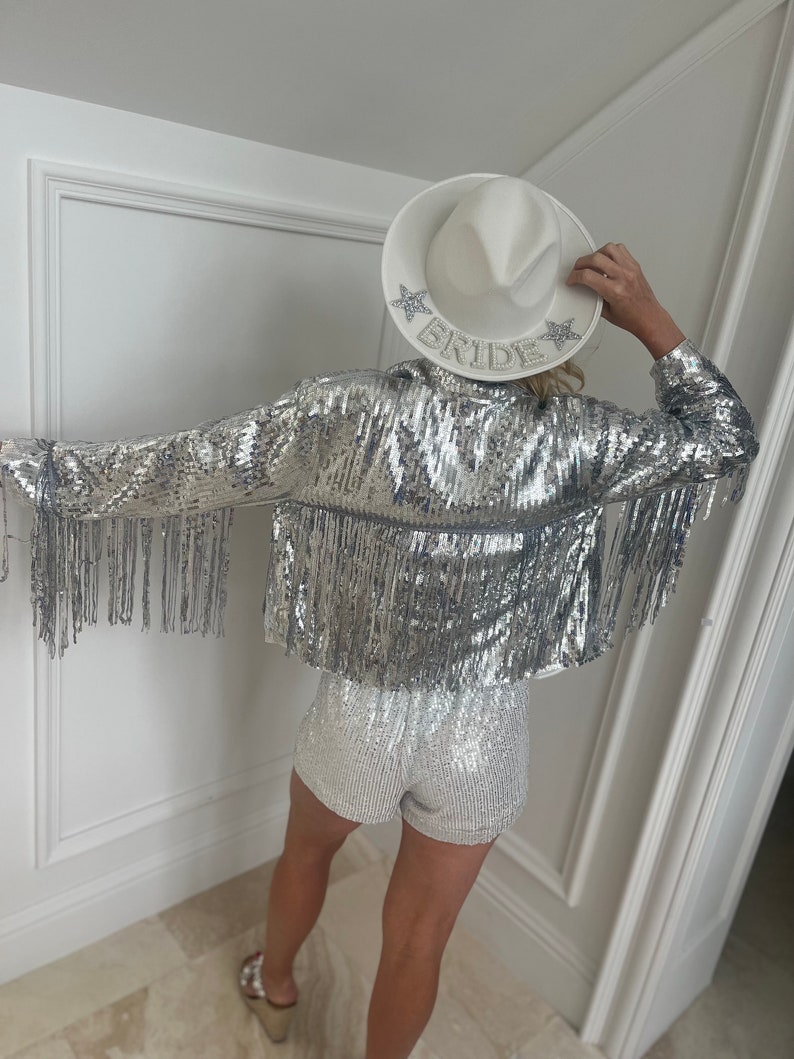 A woman in back view wearing a silver Sequin fringe cowgirl jacket with a white and silver sequin romper and a white cowgirl bride hat. The woman is posed looking up showcasing the jacket worn for bachelorette parties.