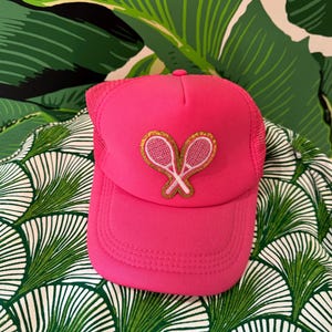 May include: A bright pink trucker hat with a mesh back and a curved brim. The hat features a gold-outlined patch of two crossed tennis rackets. The hat is set against a background of green and white patterned fabric and a green leaf-patterned wall.