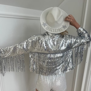 A woman in front view wearing a silver Sequin fringe cowgirl jacket with a white and silver sequin romper and a white cowgirl bride hat. The woman is posed looking down not showing her face and showcasing the jacket worn for bachelorette parties.