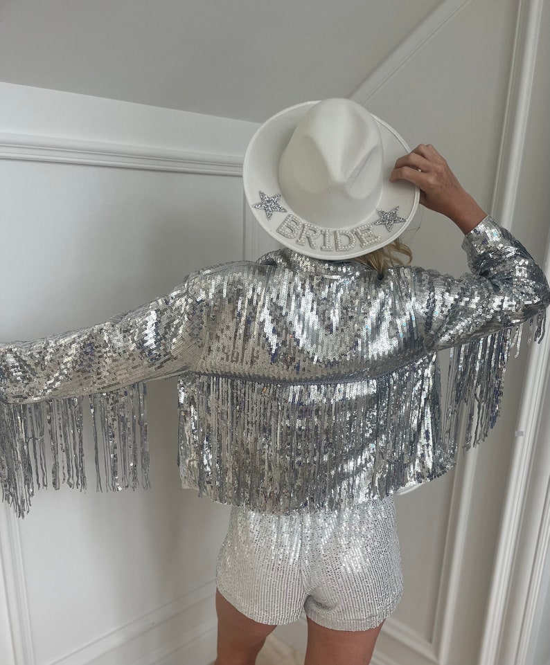 A woman in front view wearing a silver Sequin fringe cowgirl jacket with a white and silver sequin romper and a white cowgirl bride hat. The woman is posed looking down not showing her face and showcasing the jacket worn for bachelorette parties.