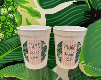 May include: Two white plastic cups with pink and green palm tree designs and the text "PALMS Social Club".