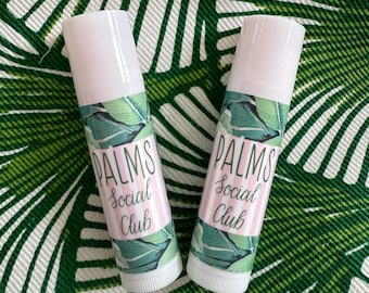 May include: Two white lip balm tubes with green and pink labels that say "PALMS Social Club". The tubes are on a green and white patterned surface.