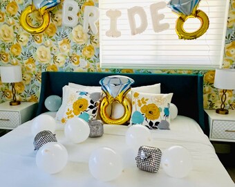 May include: A white bed with a teal headboard and white bedding. The bed is decorated with white balloons, silver balloons, and gold ring balloons. The wall behind the bed is covered in a floral wallpaper with yellow and blue flowers. The word "BRIDE" is spelled out in white letters above the bed.