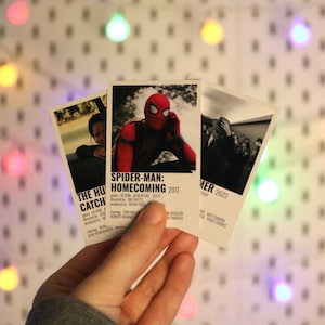 Pu&ograve; includere: Tre stampe di poster di film con Spider-Man: Homecoming, Hunger Games: La ragazza di fuoco e Summer 2023. Le stampe sono tenute in una mano su uno sfondo di luci colorate.