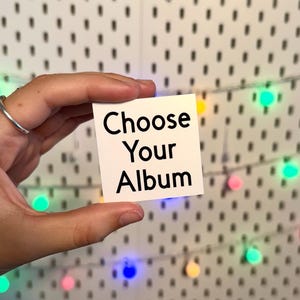 May include: A square white card with the black text Choose Your Album printed in a clean sans-serif font. The card is held against a white pegboard background featuring a string of small, colorful, glowing LED lights.