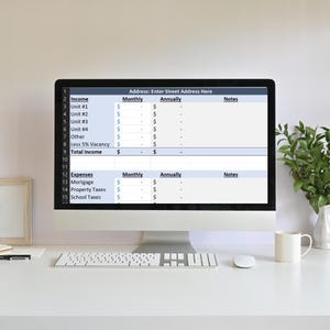 May include: A computer monitor displays a spreadsheet with financial data, including income and expenses. The screen shows columns for monthly and annual figures, with a section to enter an address. A keyboard, mouse, and coffee mug are on the white desk.