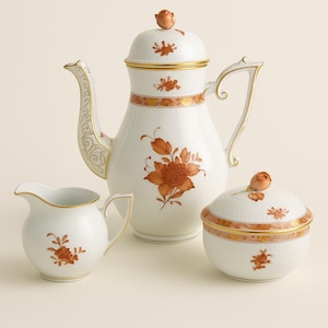May include: A white porcelain tea set with orange floral designs. The set includes a teapot, creamer, and sugar bowl. The teapot has a rose-shaped finial and gold trim. The creamer and sugar bowl also have gold trim and floral accents.