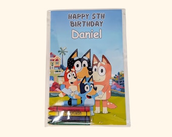 May include: A rectangular birthday card in a clear plastic sleeve featuring the text HAPPY 5TH BIRTHDAY Daniel. The card displays a colorful cartoon illustration of three blue and orange dog characters standing in front of a house, with four crayons.