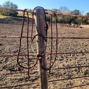 Puede incluir: Un cabestro de cuerda burdeos cuelga de un poste de madera desgastado. El cabestro está anudado y tiene un lazo. El fondo muestra una escena rural con una valla, suelo de tierra y un caballo a lo lejos.