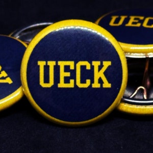 May include: Four blue and yellow round buttons with the word "UECK" printed in yellow letters.