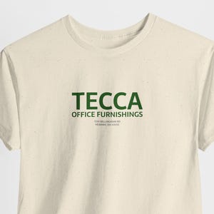 May include: A cream-colored t-shirt with the word "TECCA" in dark green block letters. Below the word is "OFFICE FURNISHINGS" in smaller green letters. The shirt also has the address "1504 BELLMEADE RD NEWARK, OH 43055".