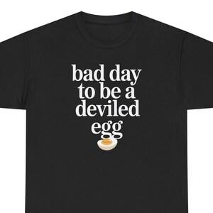 May include: Black t-shirt with the phrase "bad day to be a deviled egg" in white, with a graphic of a deviled egg. The shirt is made of a soft material and is suitable for casual wear.