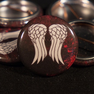 Puede incluir: Cinco botones redondos con fondo rojo y negro. El botón central presenta un gráfico blanco de alas de ángel.