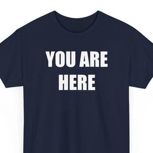 Pode incluir: Camiseta azul marinho com o texto branco "YOU ARE HERE" centralizado na frente. A camiseta de manga curta é feita de um material macio e é adequada para uso casual.