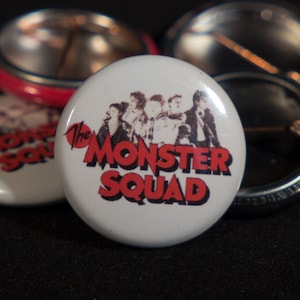 Puede incluir: Cinco pines de botón blancos con un diseño rojo y negro. El pin central presenta un grupo de cinco personas con el texto "The Monster Squad" en letras rojas.