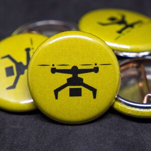May include: Four yellow round buttons with black graphic designs. One button features a drone with a package underneath. Another button features a stick figure with arms outstretched. The other two buttons are blank.