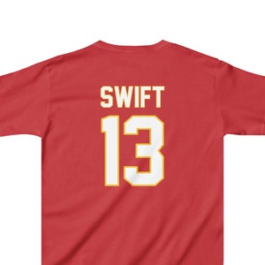 May include: Red t-shirt with the name "SWIFT" and the number "13" in white with a gold outline. The shirt is a solid red color and has short sleeves. The text is centered on the back of the shirt.