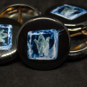 May include: Four black button pins with a white polar bear in a blue ice cube design. The polar bear is waving its arms and has a happy expression.