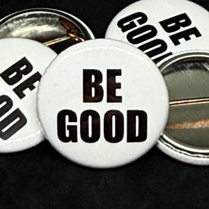 May include: A collection of white button badges with the words "BE GOOD" printed in bold, black letters. The badges have a pin backing and are arranged against a dark background. The design is simple and eye-catching.