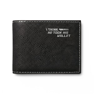 Könnte beinhalten: Ein schwarzes Portemonnaie mit zwei Fächern, einer strukturierten Oberfläche und weißen Kontrastnähten an den Rändern. Auf der Vorderseite befindet sich der Text I THINK HE TOOK HIS WALLET in einer hellgrauen, leicht abgenutzten Schrift.