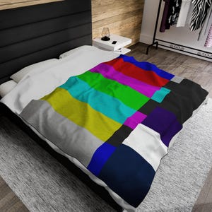 May include: A white bed with a colorful TV test pattern blanket. The blanket is made of soft fabric and features a classic TV test pattern design with stripes of red, green, blue, yellow, cyan, magenta, and black.