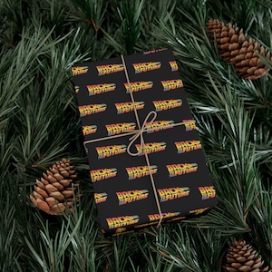 May include: A black gift wrapped in paper featuring the "Back to the Future" logo in yellow, orange, and red. The gift is tied with twine and set on a bed of green pine needles and brown pine cones.