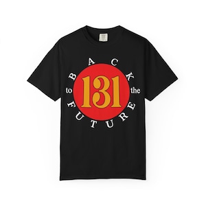 Puede incluir: Una camiseta negra de manga corta con un gráfico central que muestra un círculo rojo con el número 131 en amarillo. Las palabras BACK to the FUTURE están impresas en letras mayúsculas blancas dispuestas en círculo alrededor del rojo.
