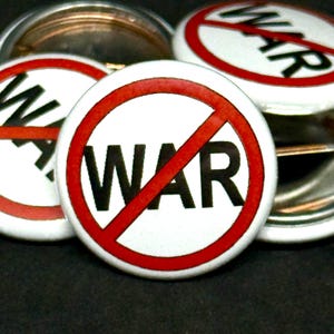 May include: A collection of white buttons with a red circle and a red diagonal line through the word "WAR" in black, conveying an anti-war message. The buttons have a metal backing and are arranged on a dark surface.