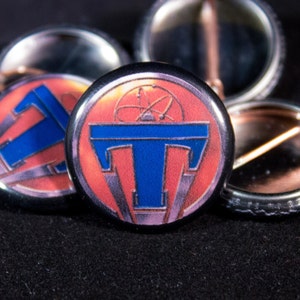 May include: A close-up of a red, white, and blue button with a stylized "T" and a circular design. The button is surrounded by other buttons.