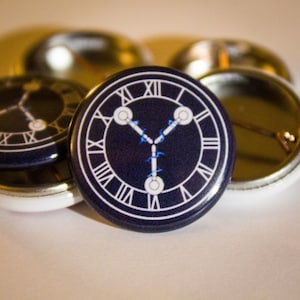 May include: A black and white button with a clock face and a stylized 'Y' shape in the center. The clock face has Roman numerals and the 'Y' shape is blue.