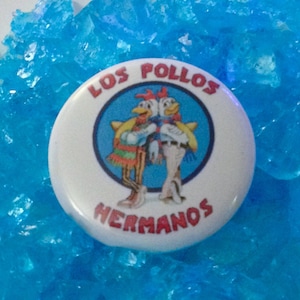 Peut inclure: Un bouton blanc avec un design bleu et rouge. Le bouton présente un logo avec deux poulets de dessin animé et le texte "Los Pollos Hermanos".