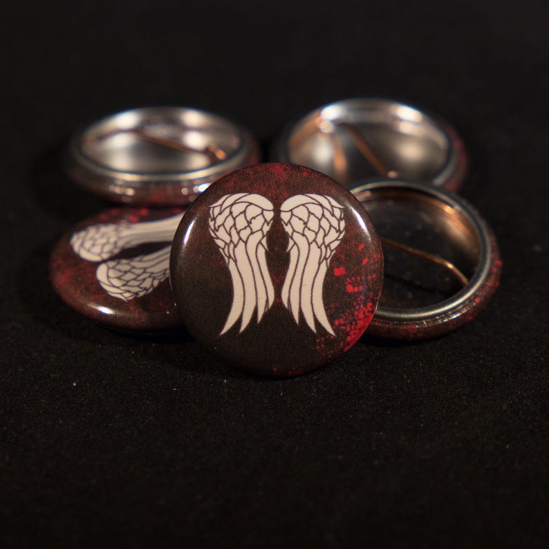 May include: Five round button pins with a white angel wing design on a dark brown background with red splatters. The wings are centered on the button and are the main focus of the design.