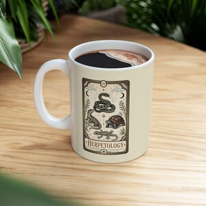 May include: A cream-coloured ceramic mug featuring a rectangular graphic design that resembles a tarot card. The illustration depicts a snake, a lizard, a turtle, and a gecko, with the text "HERPETOLOGY" printed in a decorative font at the bottom.
