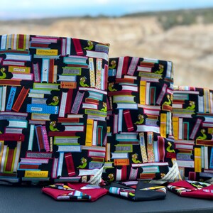 May include: Three black fabric bags with a book print design. The bags are different sizes and have a bookworm design. The bags are lined up on a dark surface.