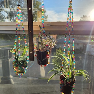 Puede incluir: Tres macetas colgantes con hilos de cuentas de colores. Cada maceta contiene una planta diferente. Las macetas están suspendidas de una barra de metal cerca de una ventana. Las cuentas son de un arcoíris de colores.