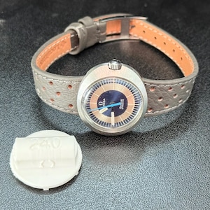 Può includere: Un orologio Omega Seamaster vintage con cassa color argento e quadrante blu e oro. L'orologio ha un cinturino in pelle grigia con un design traforato. Un componente bianco dell'orologio è accanto all'orologio.