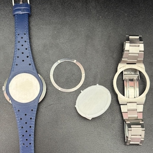 May include: A disassembled watch with a blue leather strap, a silver metal bracelet, a silver metal ring, and a white oval-shaped piece. The blue strap has perforations. The watch components are on a dark surface.