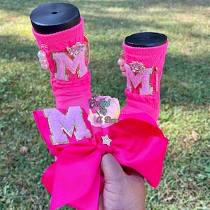 May include: Bright pink socks with the letter "M" appliqued on them, accented with gold trim and a large pink bow. The socks are decorated with pearl and rhinestone embellishments. The text "Created By A Boss" is also visible.