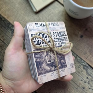 May include: A stack of rectangular cards titled Blackout Press, tied with brown twine. The top card features a vintage newspaper design with the text Strength Stands, Compassion Conquers, and an illustration of a person with a lion labeled VIII Strength.