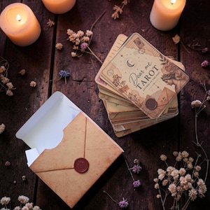 May include: A deck of Dear Me Tarot cards with a vintage aesthetic, featuring a crescent moon, botanical illustrations, and a wax seal design. The deck is accompanied by a matching tan envelope with a red wax seal closure, all arranged neatly.