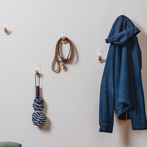May include: Four wall-mounted hooks, each featuring a white rectangular base and a cylindrical wooden peg. The hooks are arranged on a light-colored wall, holding a blue hooded jacket, a brown leather leash, and a folded blue and white striped umbrella.