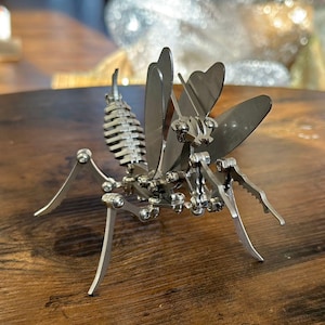 May include: A silver metal praying mantis sculpture with articulated legs and wings. The mantis is posed in a defensive stance with its front legs raised.