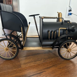 May include: A black metal model of a vintage car with gold accents. The car has a detailed design with a steering wheel, a seat, and a working engine. The car is a decorative piece and is perfect for adding a touch of vintage style to any home.